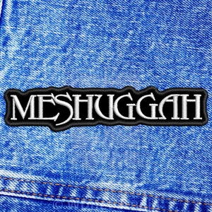 May include: A rectangular embroidered patch featuring the word MESHUGGAH in white capital letters with a black border. The patch is centred on a blue denim fabric background with visible stitching along the left side of the material.