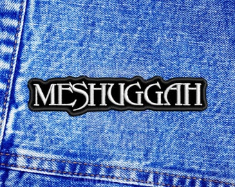 Puede incluir: Un parche rectangular bordado que presenta la palabra MESHUGGAH en letras mayúsculas blancas con un borde negro. El parche está centrado sobre un fondo de tela vaquera azul con costuras visibles a lo largo del lado izquierdo.