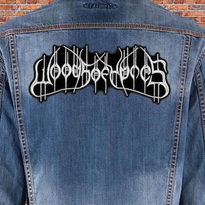 May include: A black and white embroidered patch with the text "Woods of Yore" on a denim jacket.