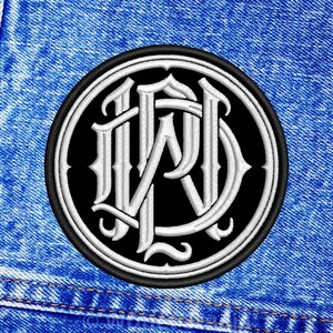 Op de afbeelding: Ronde geborduurde patch met een wit monogram van de letters "WRD" in gotische stijl op een zwarte achtergrond. De patch heeft een zwarte rand en wordt weergegeven op een blauwe denim stof.
