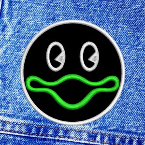 May include: Black and white embroidered patch with a green mouth and two white eyes. The patch is sewn onto a blue denim background.