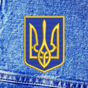 Może przedstawiać: Złota haftowana naszywka z ukraińskim herbem, trójzębem, na niebieskim tle. Naszywka jest przyszyta do niebieskiego materiału dżinsowego.