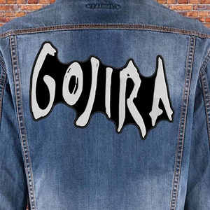 Peut inclure: Un écusson brodé noir et blanc avec le mot "GOJIRA" dans une police de caractères stylisée. L'écusson est cousu sur une veste en jean bleue.