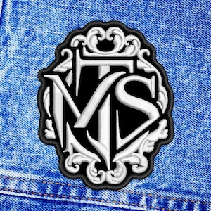 May include: A black and white embroidered patch featuring the letters "M.S." within an ornate frame. The patch is displayed against a blue denim backdrop.