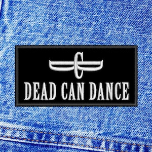 Può includere: Una toppa rettangolare nera con la scritta ricamata "DEAD CAN DANCE" in bianco, insieme a un emblema bianco stilizzato. La toppa è posta su uno sfondo di denim blu, con cuciture visibili.