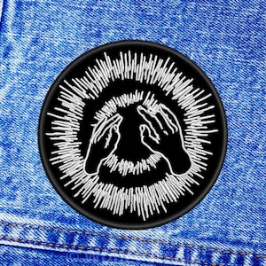 May include: Black and white embroidered patch featuring two hands reaching up towards a circle of white lines. The patch is sewn onto a blue denim background.