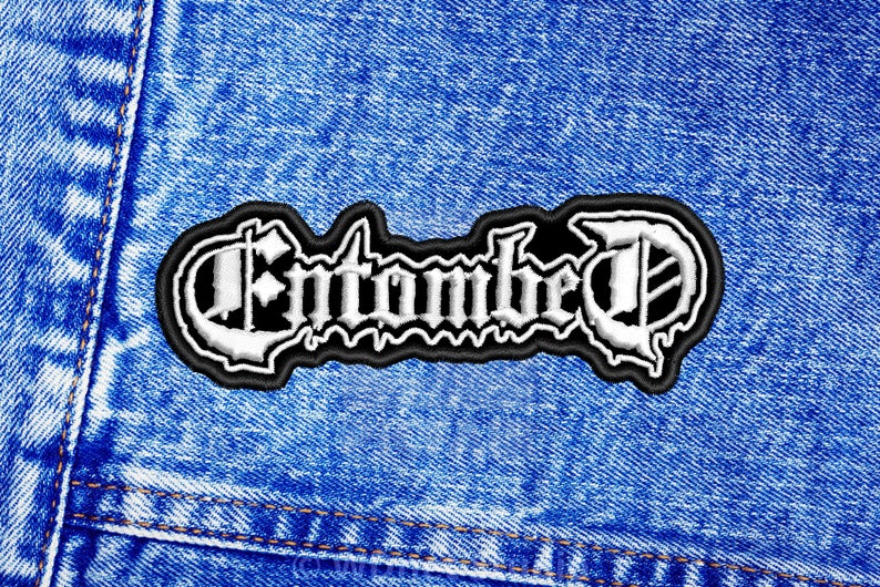 K&ouml;nnte beinhalten: Wei&szlig;er gestickter Aufn&auml;her mit dem Wort "Entombed" in gotischer Schrift auf schwarzem Hintergrund. Der Aufn&auml;her ist auf eine blaue Jeansjacke gen&auml;ht.