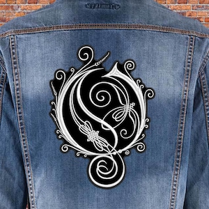 May include: A blue denim jacket featuring a large, circular embroidered patch on the back. The patch is black with intricate white filigree and scrollwork designs forming a stylized letter O, surrounded by decorative swirling patterns.