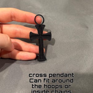 Może przedstawiać: Czarny wisiorek w kształcie krzyża z okrągłym oczkiem u g&oacute;ry. Wisiorek ma teksturowaną powierzchnię i zawiera tekst: cross pendant Can fit around the hoops or inside chains. Projekt jest symetryczny z rozszerzającymi się końcami ramion krzyża.