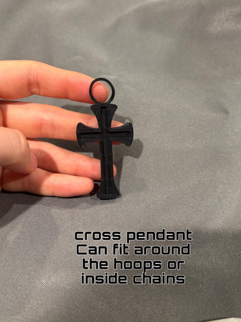 Może przedstawiać: Czarny wisiorek w kształcie krzyża z okrągłym oczkiem u g&oacute;ry. Wisiorek ma teksturowaną powierzchnię i zawiera tekst: cross pendant Can fit around the hoops or inside chains. Projekt jest symetryczny z rozszerzającymi się końcami ramion krzyża.