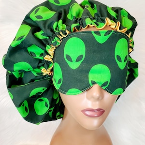 May include: A dark green sleep bonnet and eye mask set featuring a repeating pattern of green alien faces. The bonnet has a ruffled edge with a gold satin trim. The eye mask also has a gold satin trim.