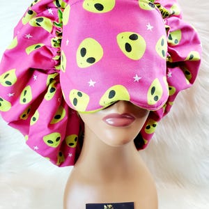 May include: Pink satin sleep cap with a matching sleep mask. Both feature a green alien face pattern with white stars. The sleep cap has a gathered elastic band. The sleep mask has a black elastic band. The text "Eve's Satin Crown Co. Luxury Essentials & Unique Finds" is on a white card in the lower right corner of the image.