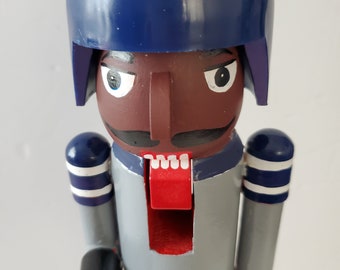 May include: A wooden nutcracker with a blue helmet, gray uniform, and white stripes. The nutcracker has a brown face with a black mustache and a red mouth.