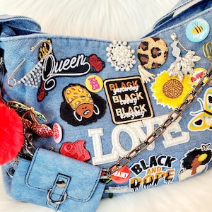 May include: A light blue denim handbag adorned with patches, pins, and a chain strap. Patches include the word "LOVE", a sunflower, a butterfly, and the text "BLACK yesterday BLACK today BLACK everyday". A small denim purse and a red pom-pom are attached.