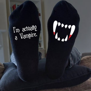 May include: Black socks with white text reading "I'm actually a Vampire." The other sock displays a white vampire teeth design with red accents. The socks are worn on a person's feet.