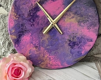 May include: A round clock with a pink, purple, and gold abstract design. The clock hands are gold.