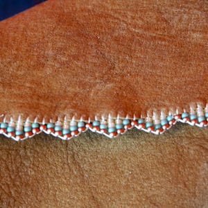 May include: Brown leather with a scalloped edge trimmed with a beaded border. The beads are light blue, white, and brown.