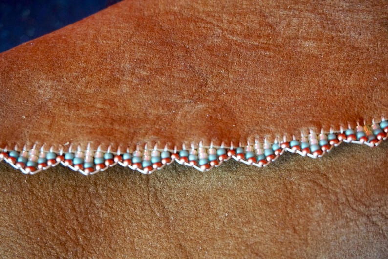 May include: Brown leather with a scalloped edge trimmed with a beaded border. The beads are light blue, white, and brown.