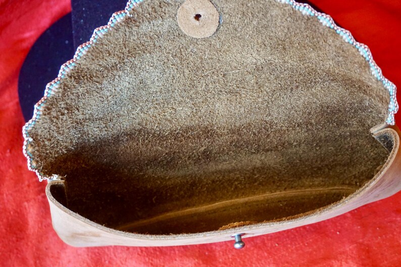 May include: A brown leather clutch bag with a scalloped edge and a beaded trim. The bag is open and the inside is lined with brown suede.