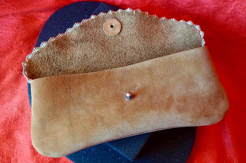 May include: A brown leather clutch bag with a scalloped edge and a snap closure. The bag is open and the inside is lined with a brown fabric.