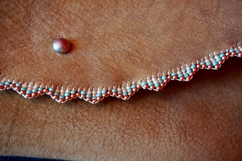 May include: Close-up of a brown leather purse with a scalloped edge trimmed with a beaded border of copper and light blue beads.