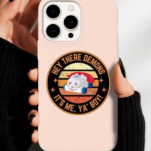 Puede incluir: Funda de teléfono rosa claro con un diseño de teléfono de dibujos animados retro y el texto "HEY THERE DEMONS IT'S ME, YA BOY!" en un diseño circular. La funda tiene un acabado brillante y la sostiene una persona.