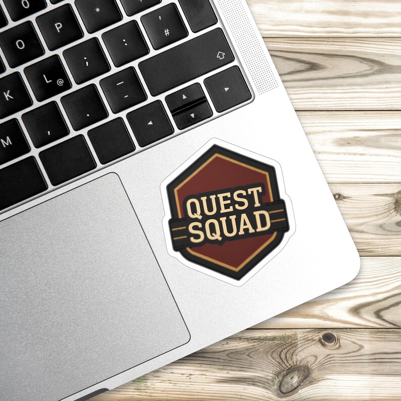 Puede incluir: Una pegatina con el texto "QUEST SQUAD" en negrita. La pegatina tiene forma hexagonal, marr&oacute;n y negra, con un borde dorado. Est&aacute; colocada sobre un teclado de port&aacute;til y una superficie de madera.