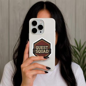 Puede incluir: Una funda de tel&eacute;fono blanca con una pegatina que dice "QUEST SQUAD" en letras doradas, sobre un dise&ntilde;o geom&eacute;trico marr&oacute;n y negro. La funda del tel&eacute;fono se sostiene frente a la cara de una persona.