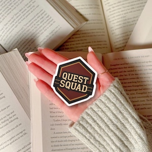 Puede incluir: Una pegatina Quest Squad marr&oacute;n y negra con un dise&ntilde;o geom&eacute;trico. La pegatina tiene un fondo marr&oacute;n oscuro con las palabras "QUEST SQUAD" en blanco y dorado. La pegatina se sostiene sobre libros abiertos.