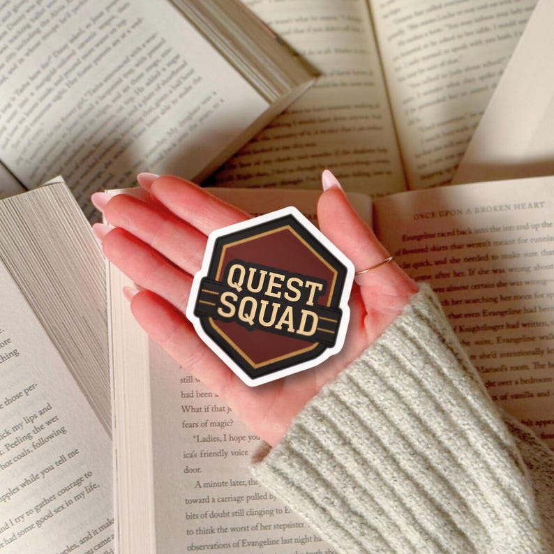 Puede incluir: Una pegatina Quest Squad marr&oacute;n y negra con un dise&ntilde;o geom&eacute;trico. La pegatina tiene un fondo marr&oacute;n oscuro con las palabras "QUEST SQUAD" en blanco y dorado. La pegatina se sostiene sobre libros abiertos.