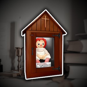 May include: A sticker featuring a doll inside a wooden cabinet with a cross on top. The doll has red hair and is wearing a white dress and red-striped socks. A sign reads "WARNING, POSITIVELY DO NOT".