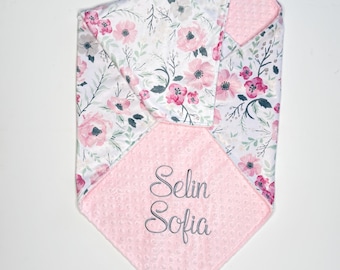 May include: A baby blanket with a floral pattern of pink flowers and green leaves on a white background. The blanket has a pink, textured square with the embroidered name "Selin Sofia".