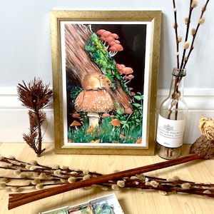 May include: A framed art print with a detailed illustration of a mouse on a mushroom, with other mushrooms and a log. The print is in a gold frame. Additional items include a watercolour palette, a brush, and decorative branches.