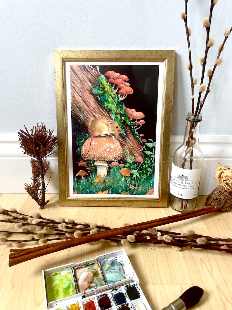 May include: A framed art print with a detailed illustration of a mouse on a mushroom, with other mushrooms and a log. The print is in a gold frame. Additional items include a watercolour palette, a brush, and decorative branches.