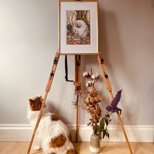 May include: A white rabbit painting on an easel with a wooden frame. The painting is set against a light grey wall. A white and brown cat sits on the floor looking at the painting. A vase with dried flowers sits on the floor in front of the easel.