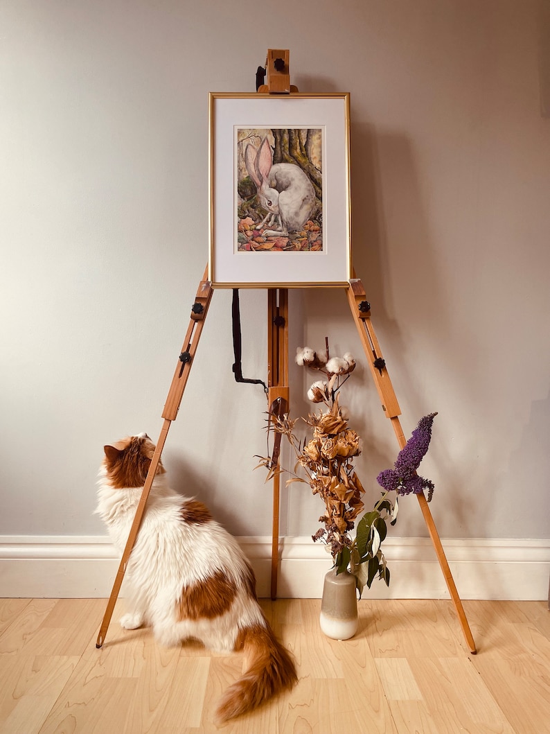 May include: A white rabbit painting on an easel with a wooden frame. The painting is set against a light grey wall. A white and brown cat sits on the floor looking at the painting. A vase with dried flowers sits on the floor in front of the easel.
