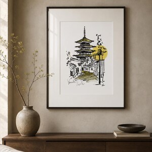 Op de afbeelding: Een ingelijste kunstdruk met een zwarte inkttekening van een pagode met meerdere verdiepingen en omliggende gebouwen. De illustratie bevat een geel cirkelvormig accent en handgeschreven tekst linksonder, alles in een dunne zwarte lijst.