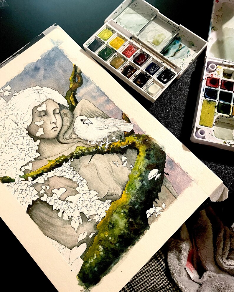 Op de afbeelding: Aquarel schilderij in uitvoering, met een serene figuur met gesloten ogen, een witte duif en een mosachtige tak. Aquarelverfpaletten met diverse kleuren omringen het kunstwerk, wat wijst op een werkruimte van een kunstenaar.
