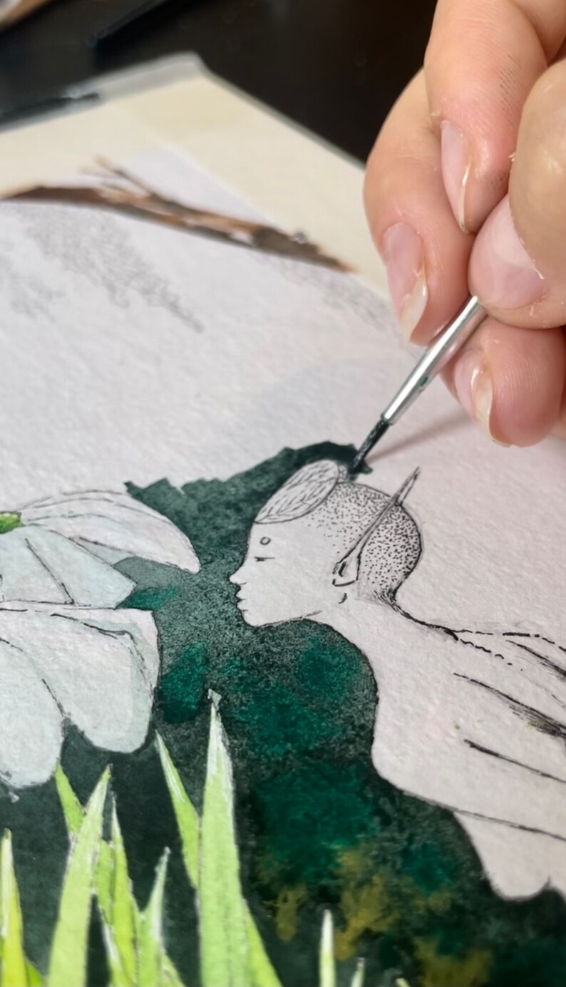 May include: Watercolor painting in progress, depicting a serene scene with a figure and stylized flora. The figure has a detailed, dotted design on the head and ear. The artist's hand holds a brush, adding details to the artwork. The color palette includes white, green, and black.