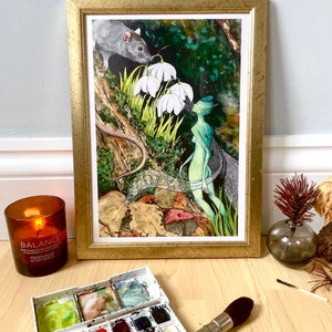 May include: A framed watercolor painting with a gold frame depicts a fairy, a rat, and white flowers in a forest setting. A lit candle, a watercolor palette, and a paintbrush are in the foreground.