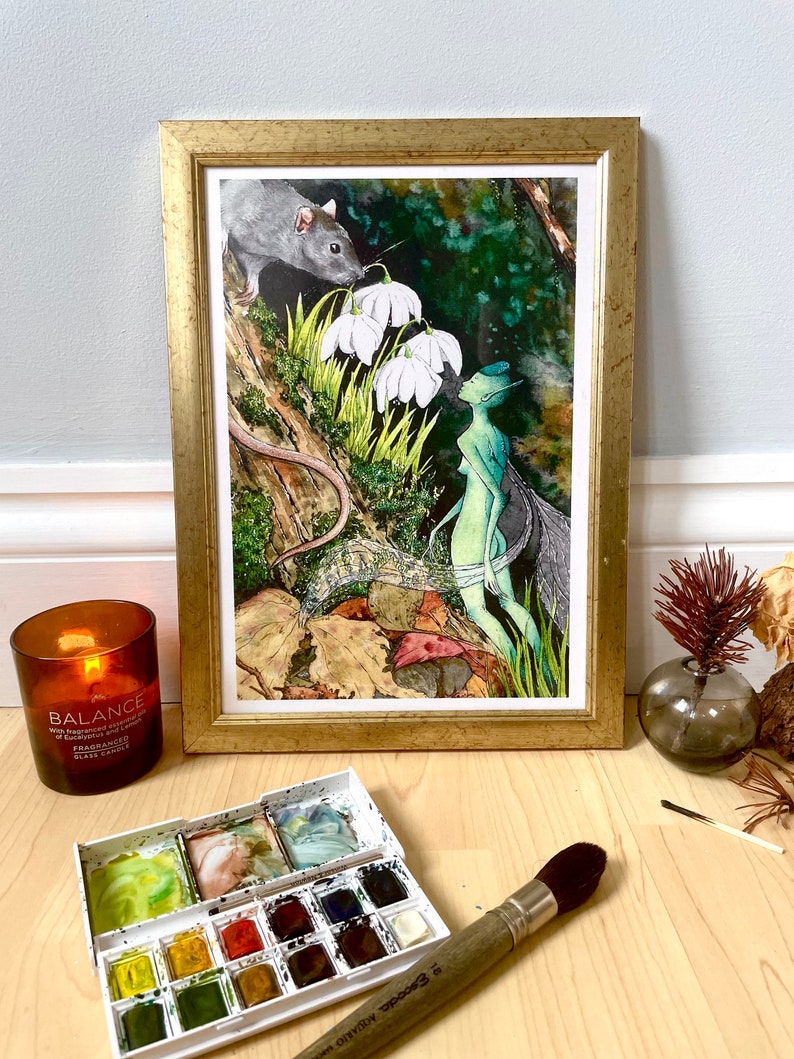 May include: A framed watercolor painting with a gold frame depicts a fairy, a rat, and white flowers in a forest setting. A lit candle, a watercolor palette, and a paintbrush are in the foreground.