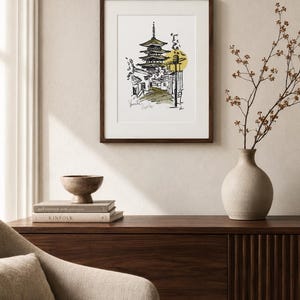 Puede incluir: Una l&aacute;mina de arte enmarcada que presenta un boceto en tinta negra de una pagoda japonesa y una escena callejera con un acento circular amarillo. El dibujo incluye texto escrito a mano en la parte inferior y se muestra en un marco de madera oscura.