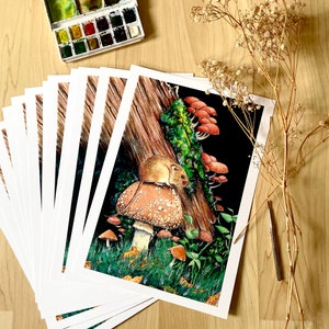 May include: A collection of art prints showcasing a detailed watercolour illustration of a mouse on a mushroom, with other fungi and foliage. A watercolour paint set and dried flowers are on a wooden surface.