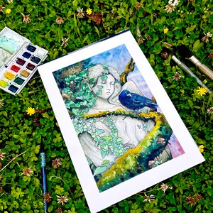 Op de afbeelding: Een aquarel schilderij van een serene figuur met gesloten ogen, versierd met klimop en een blauwe vogel, tegen een achtergrond van een blauwe lucht. Het kunstwerk wordt gepresenteerd op een witte passe-partout, omringd door kunstbenodigdheden en groen.