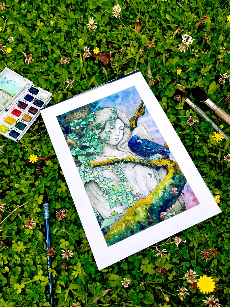 Op de afbeelding: Een aquarel schilderij van een serene figuur met gesloten ogen, versierd met klimop en een blauwe vogel, tegen een achtergrond van een blauwe lucht. Het kunstwerk wordt gepresenteerd op een witte passe-partout, omringd door kunstbenodigdheden en groen.