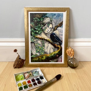 Op de afbeelding: Een ingelijste kunstdruk met een gouden lijst toont een slapende figuur versierd met klimop, met een raaf in de buurt. Een aquarelverfset en een penseel staan op de voorgrond, wat wijst op een kunstthema. Het kleurenpalet omvat groen, blauw en goud.