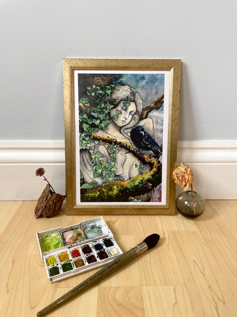 Op de afbeelding: Een ingelijste kunstdruk met een gouden lijst toont een slapende figuur versierd met klimop, met een raaf in de buurt. Een aquarelverfset en een penseel staan op de voorgrond, wat wijst op een kunstthema. Het kleurenpalet omvat groen, blauw en goud.