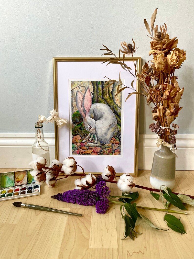 May include: A framed watercolor painting of a white rabbit with pink ears sitting in a forest setting. The painting is surrounded by dried flowers, cotton bolls, and a watercolor paint set.