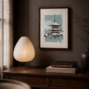 May include: A framed wall art print featuring a multi-tiered pagoda structure in dark red and white against a light blue background with a large pale moon. The artwork is displayed in a dark wood frame with a white mat border, hanging on a dark wall.