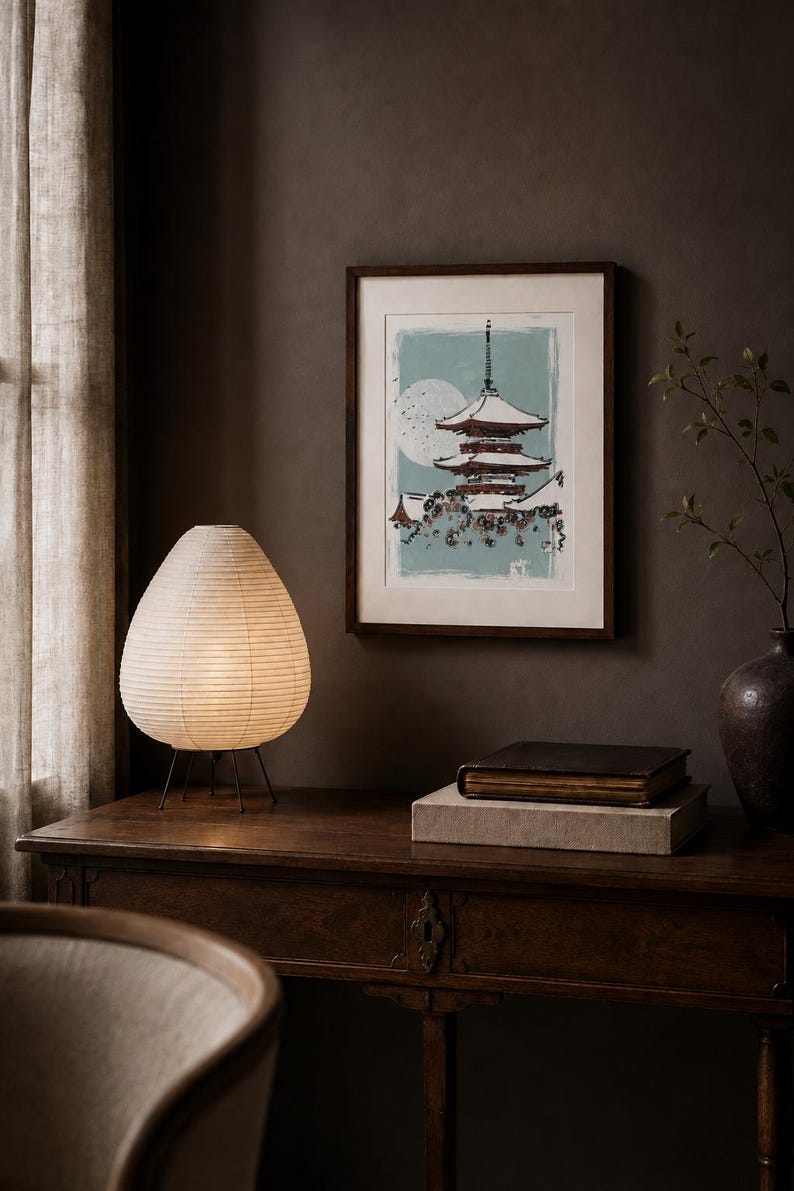May include: A framed wall art print featuring a multi-tiered pagoda structure in dark red and white against a light blue background with a large pale moon. The artwork is displayed in a dark wood frame with a white mat border, hanging on a dark wall.